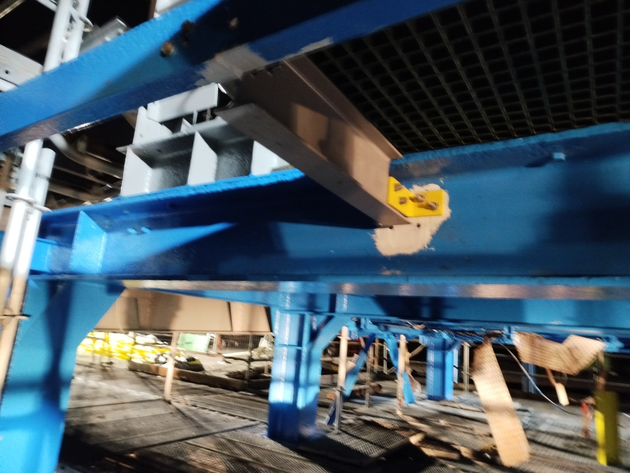 Niebieska konstrukcja stalowa w trakcie malowania, widoczne elementy rusztowania i kratownica sufitowa. Fragment konstrukcji z żółtą naklejką i śladami szpachlowania.