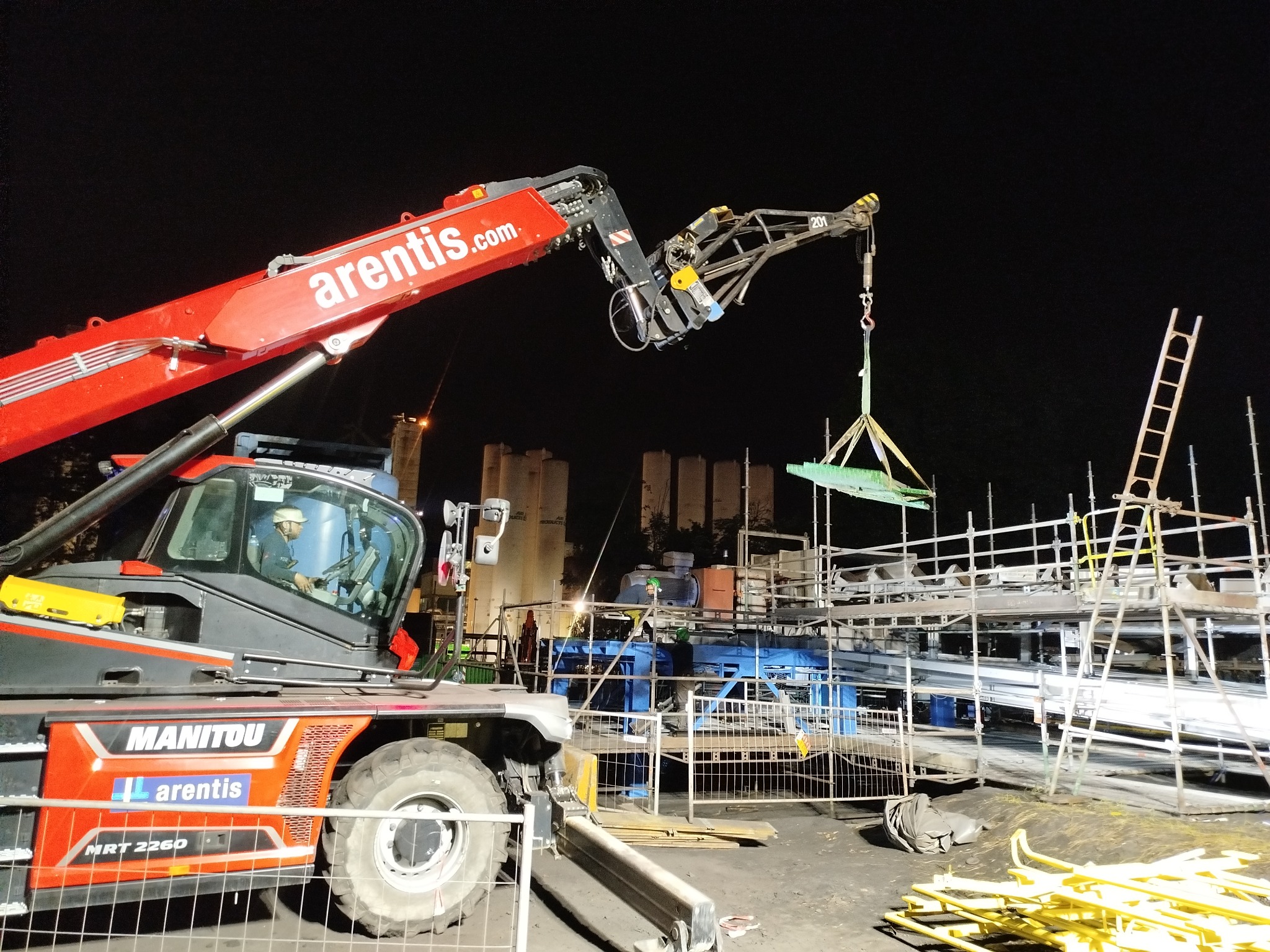 Czerwony dźwig Manitou podnosi zieloną płytę na budowie nocą, widoczne rusztowania i pracownik w kabinie.