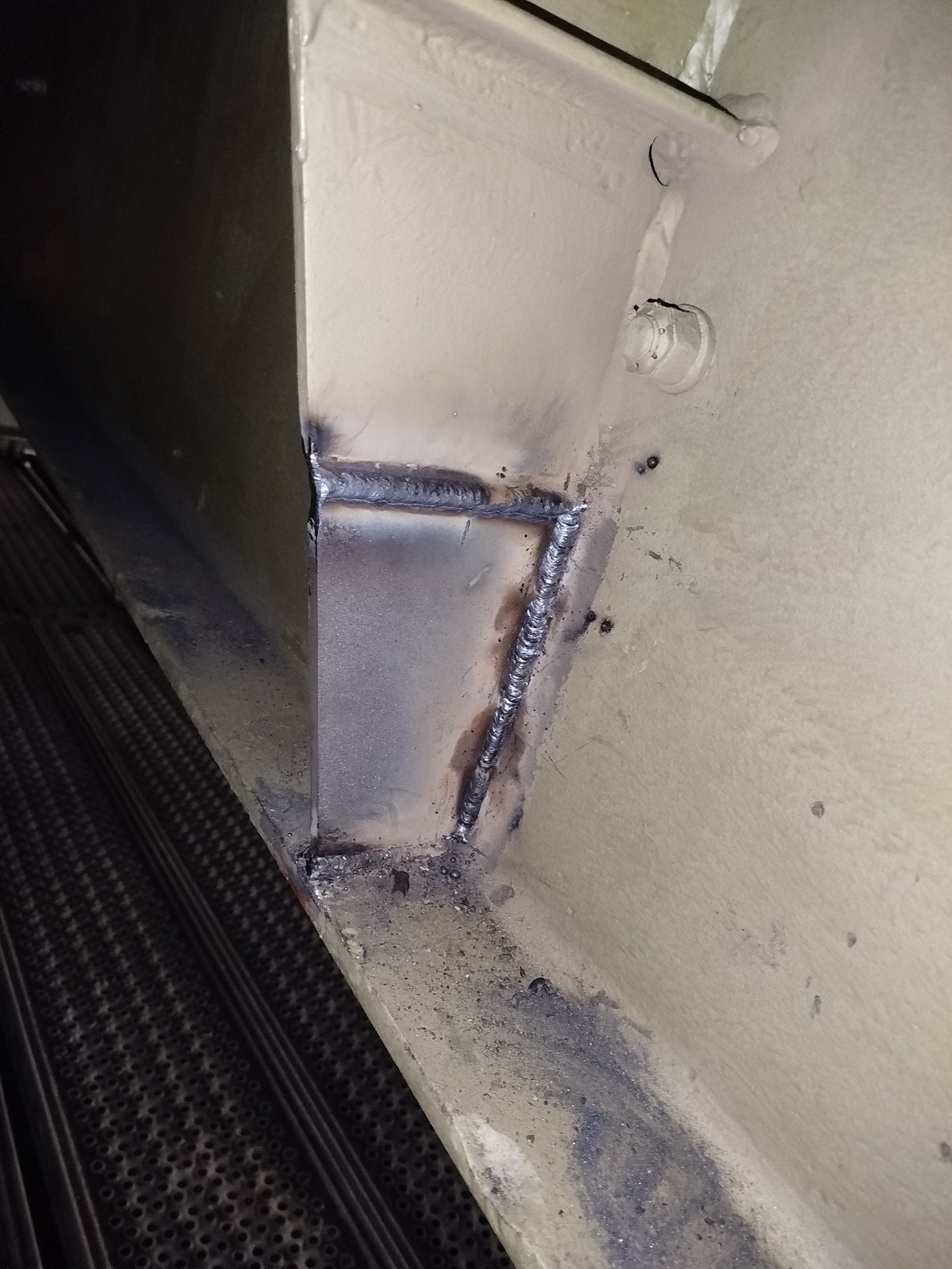 Detale spawu na beżowej konstrukcji stalowej. Widoczne złącza, ślady po spawaniu i fragment perforowanej powierzchni w tle. Niewyraźne oświetlenie.