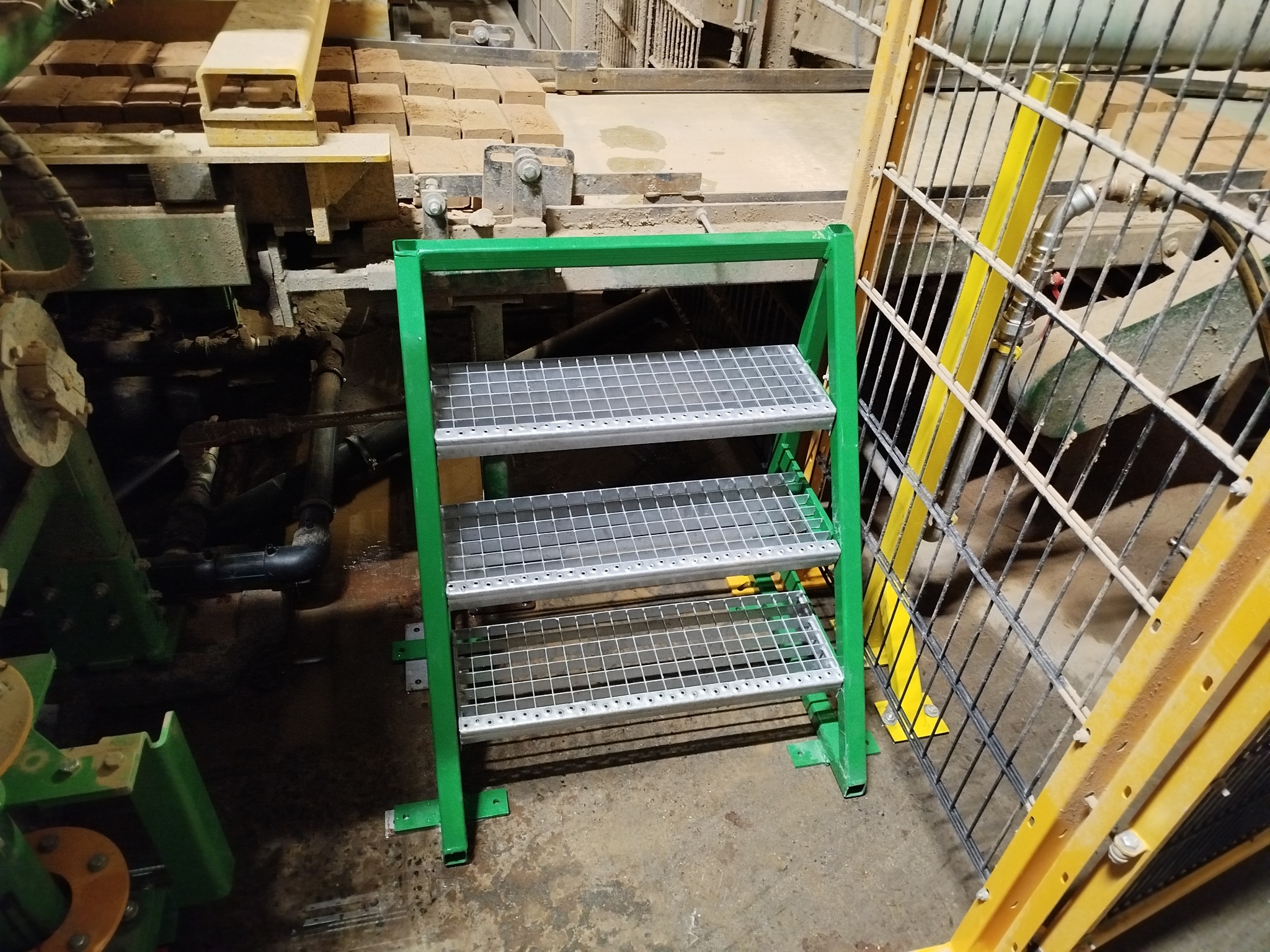 Metalowe schodki z kratką, pomalowane na zielono, stoją w przestrzeni przemysłowej. W tle widoczne maszyny i elementy konstrukcyjne.