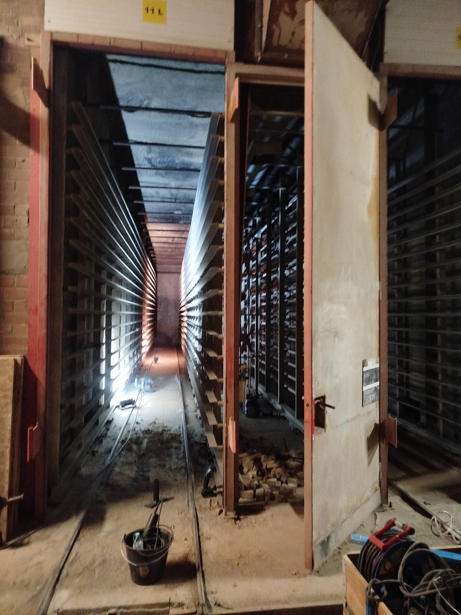 Wnętrze komory suszarniczej z metalowymi regałami, widoczne narzędzia i resztki materiałów budowlanych, oświetlenie punktowe w głębi. Drzwi komory otwarte.