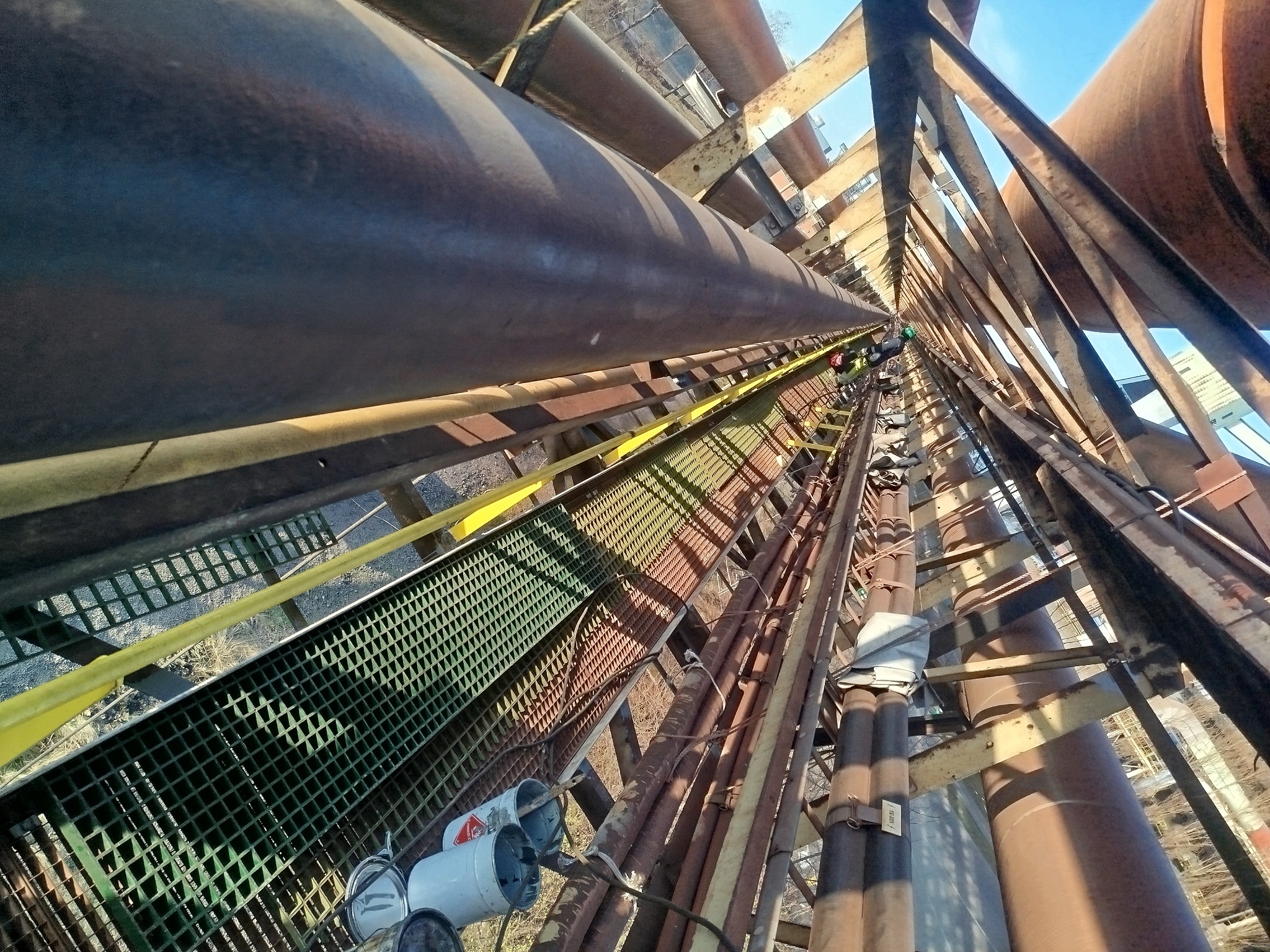 Widok z dołu na metalową konstrukcję z rurami i kratownicami, z pracownikami na górze. Perspektywa z dołu podkreśla złożoność i wysokość instalacji.