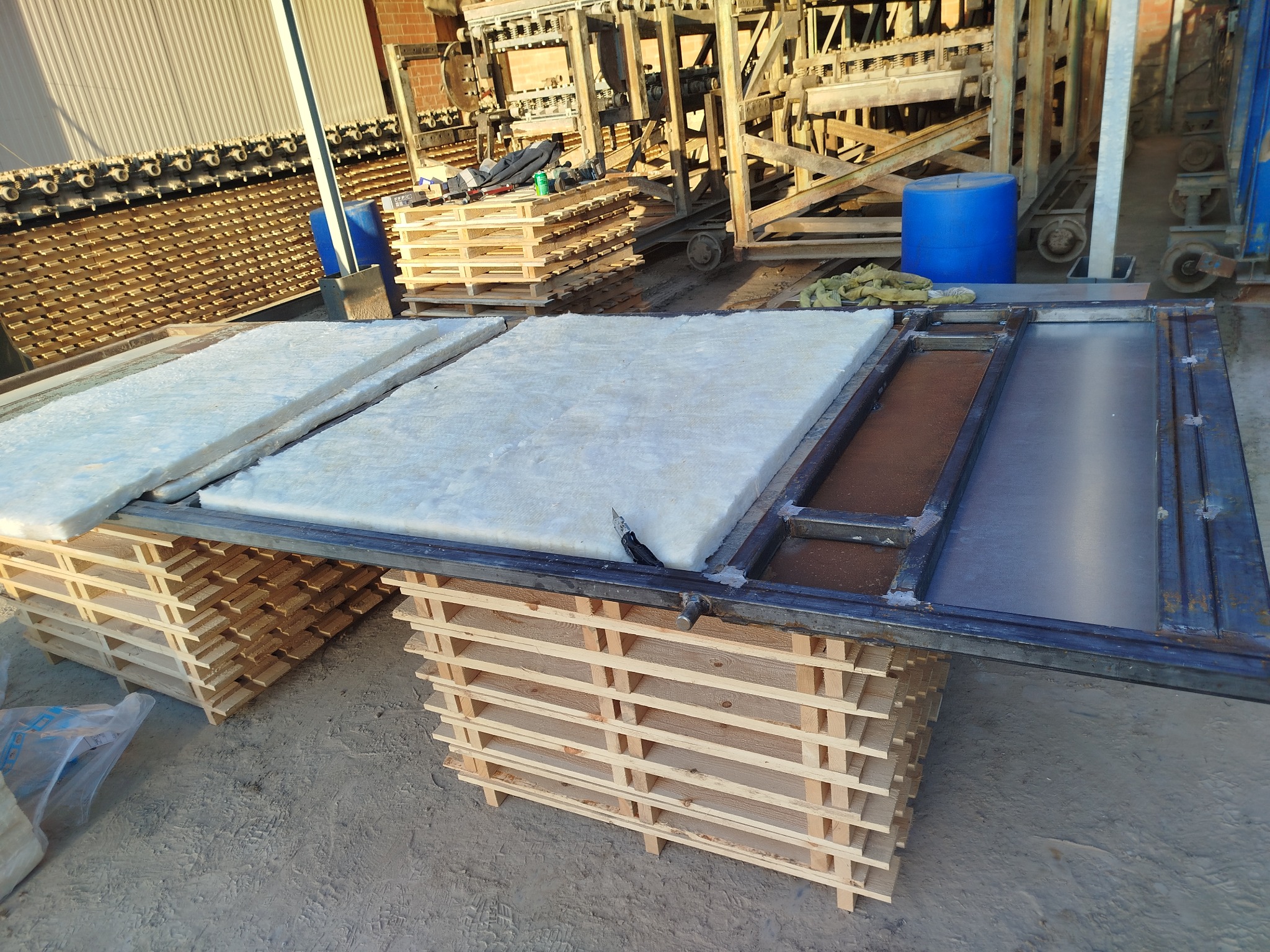 Metalowa rama z wypełnieniem z wełny mineralnej i blachy, ułożona na paletach. Widoczne narzędzia i elementy konstrukcyjne w tle.