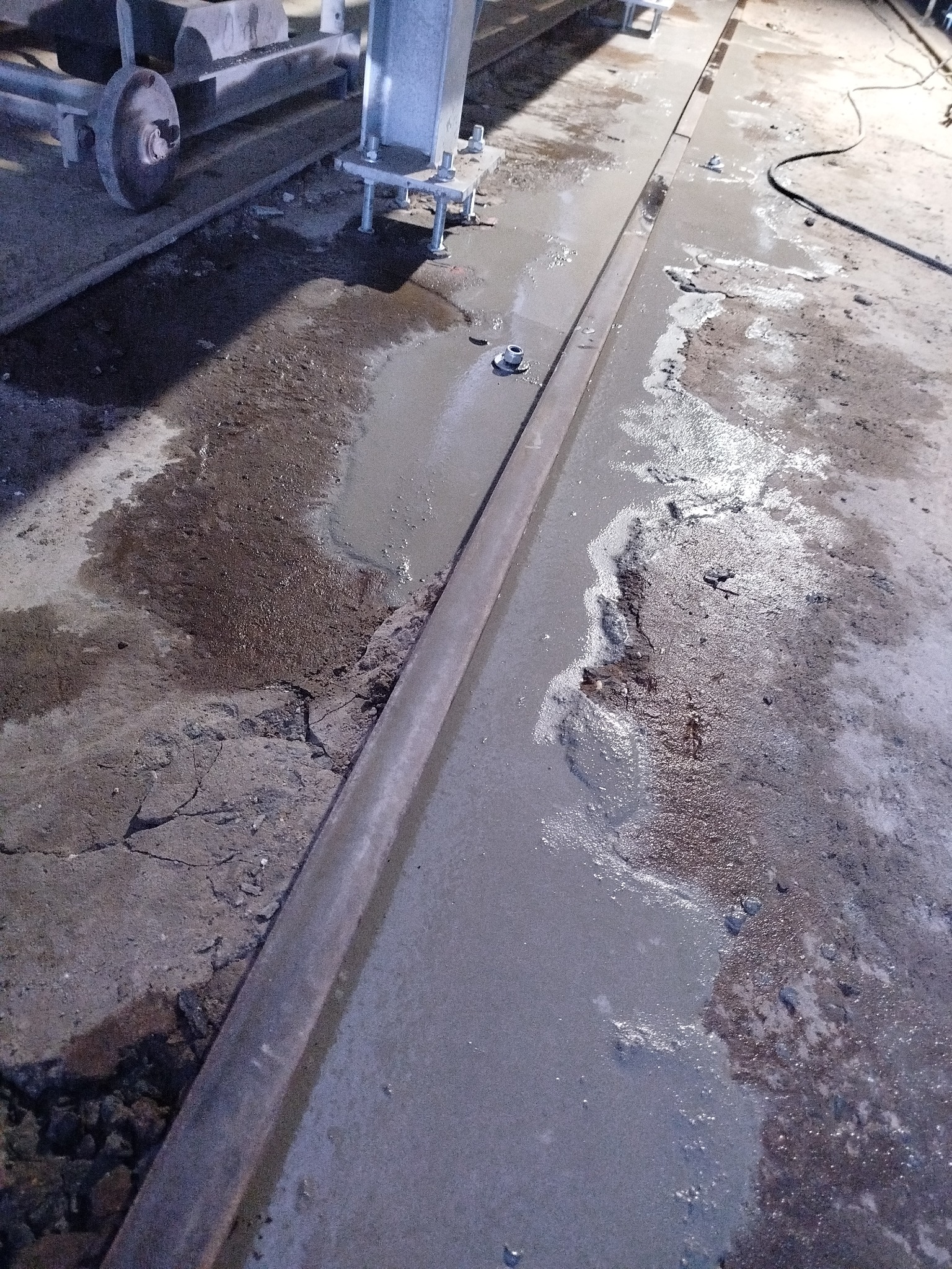 Mokry beton na podłodze, z widocznymi elementami konstrukcyjnymi i fragmentami gruzu. Ślady naprawy lub wylewki betonu na nierównej powierzchni.