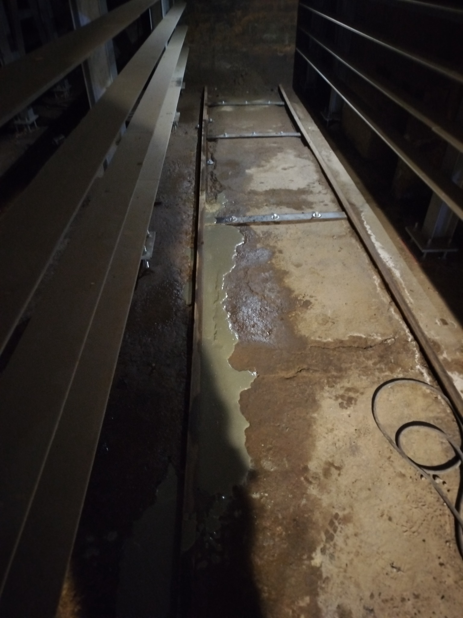 Wąska przestrzeń z betonową podłogą w trakcie prac. Widoczne metalowe elementy konstrukcyjne i ślady wilgoci. Ujęcie z dołu, ciemne otoczenie.