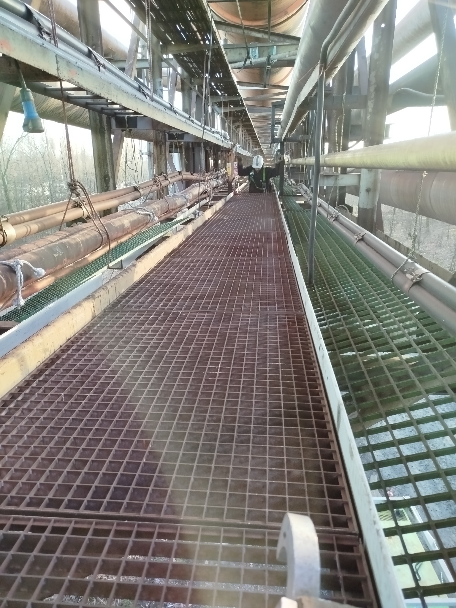 Przemysłowy pomost z metalową kratą, rurociągi i pracownik w kasku w oddali. Widok z dołu, oświetlenie dzienne.