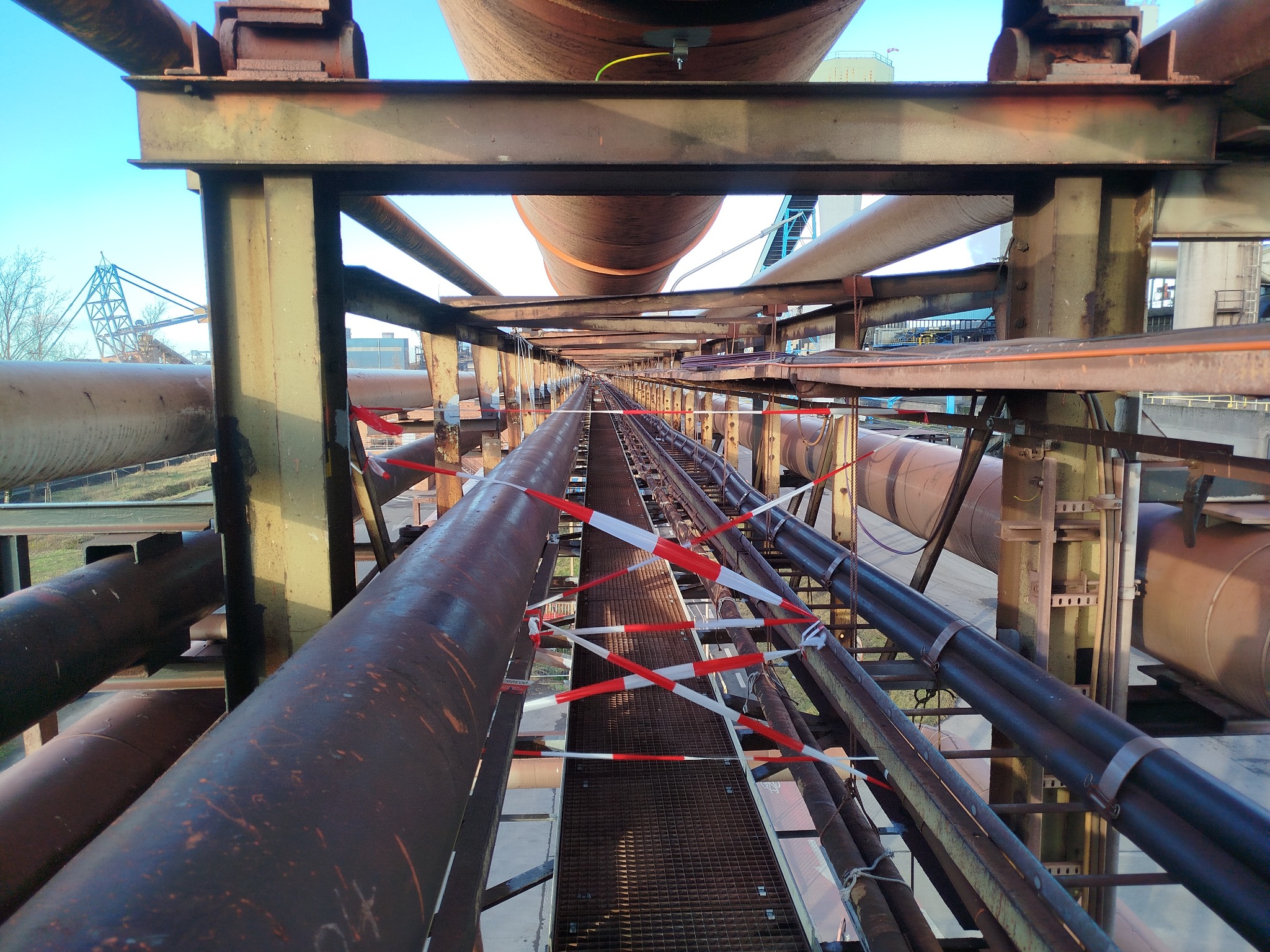 Metalowa konstrukcja z rurami i kładką, zabezpieczona taśmą ostrzegawczą. Widok w perspektywie z dołu, z elementami infrastruktury przemysłowej w tle.