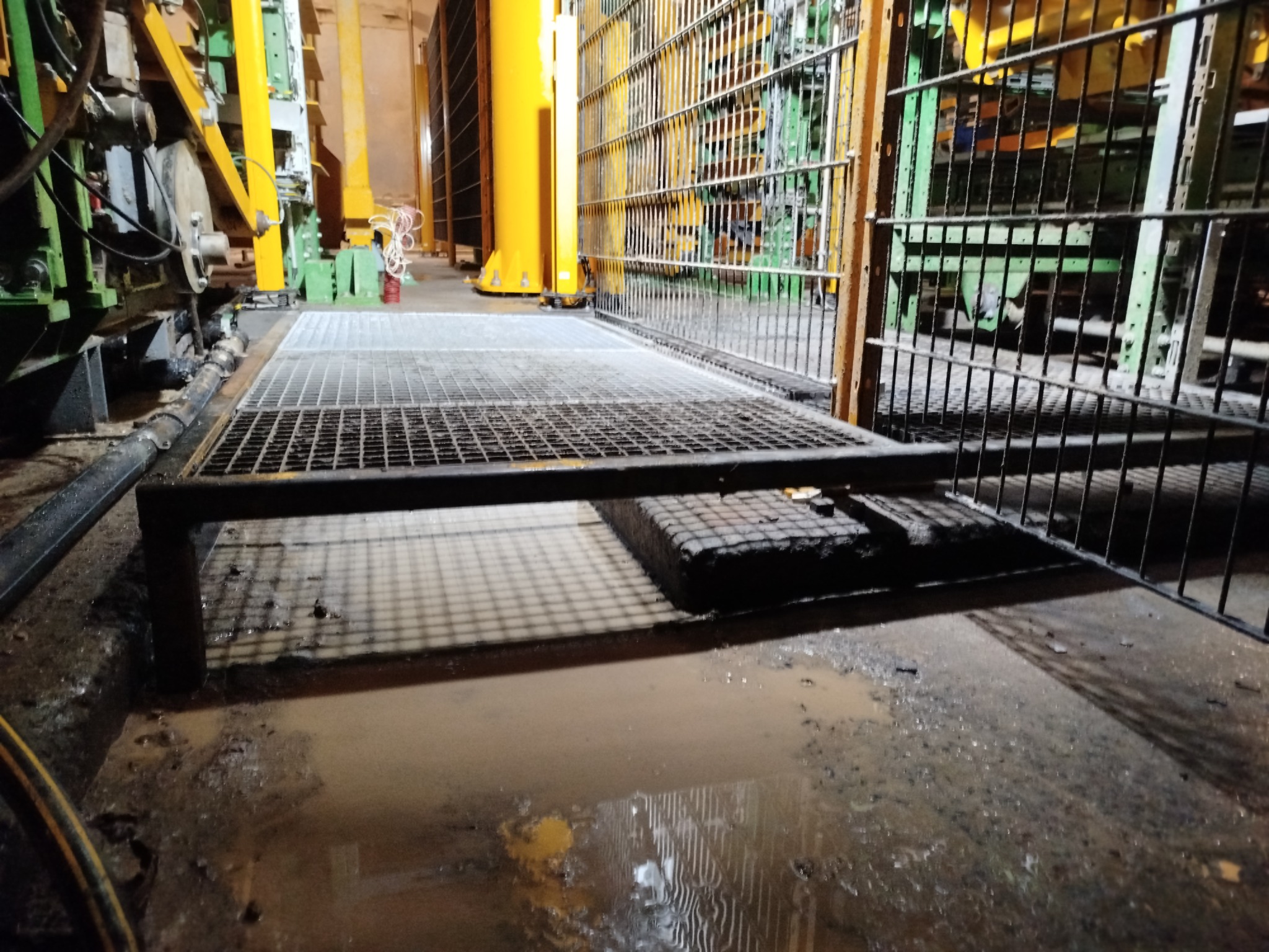 Metalowa krata podestowa w hali przemysłowej z żółtymi i zielonymi elementami konstrukcyjnymi w tle, widoczna kałuża na posadzce.