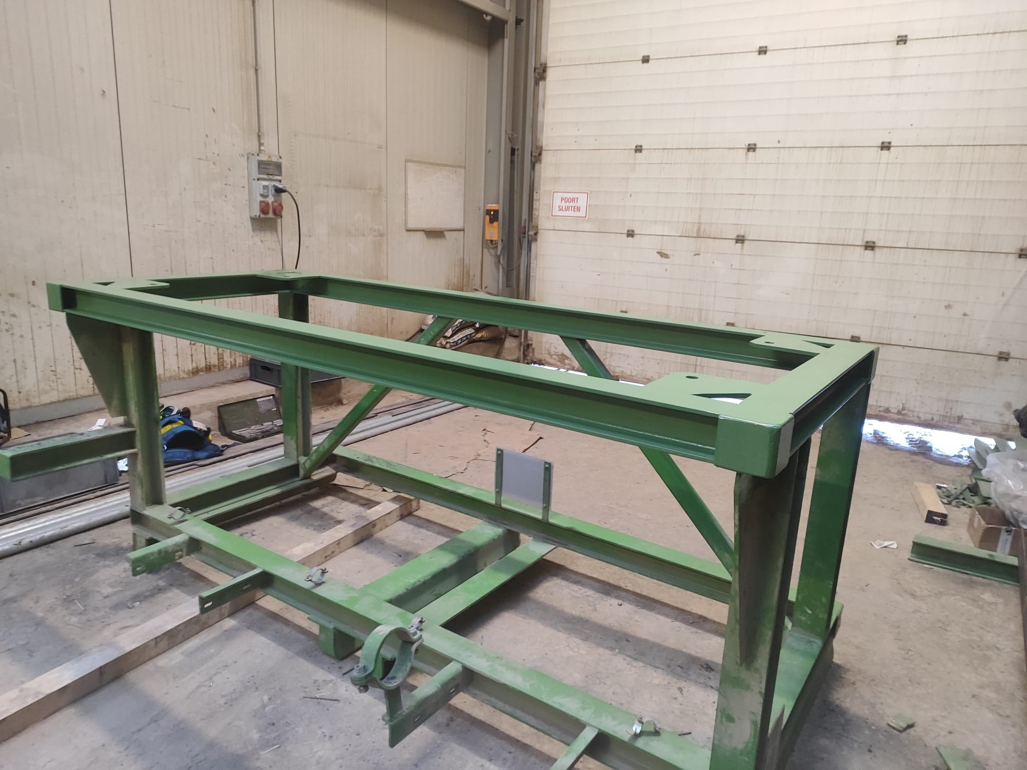 Zielona, metalowa konstrukcja spawana na hali produkcyjnej. Widoczne ślady obróbki i niedoskonałości malarskie. W tle brama garażowa i instalacje elektryczne.