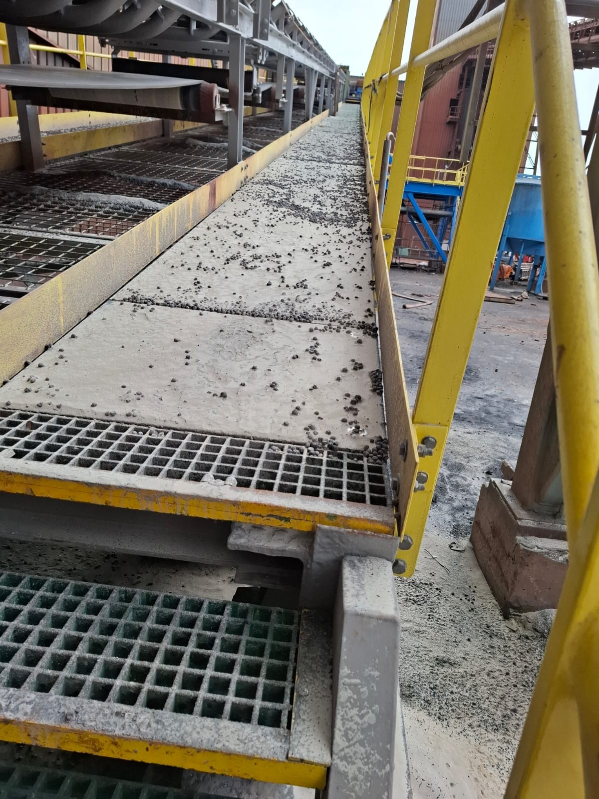 Metalowa konstrukcja z żółtymi barierkami, pokryta pyłem i drobnymi grudkami, prawdopodobnie w hali przemysłowej.