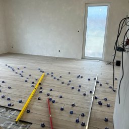 VertoAir - Układanie płytek podłogowych z użyciem poziomnic i klipsów w jasnym pomieszczeniu. Widoczny fragment ogrzewania podłogowego. Praca w toku, surowy stan ścian.