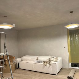 VertoAir - Wnętrze w trakcie wykończenia: drabina, sofa, lampy sufitowe. Ściany po gładziowaniu, okno zabezpieczone folią. Widoczne kartony i zwinięty dywan.