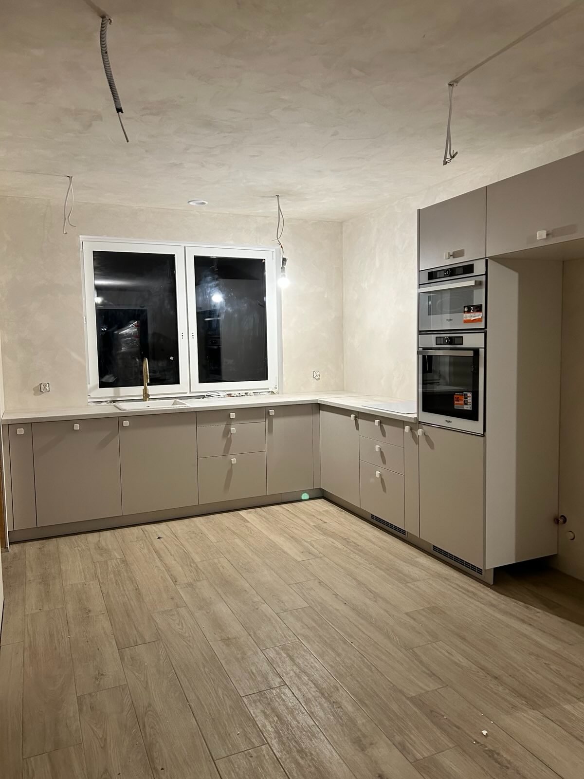 Jasna kuchnia z drewnianą podłogą i nowoczesną zabudową. Widoczne niedokończone prace elektryczne na suficie i ścianach. Minimalistyczny design.