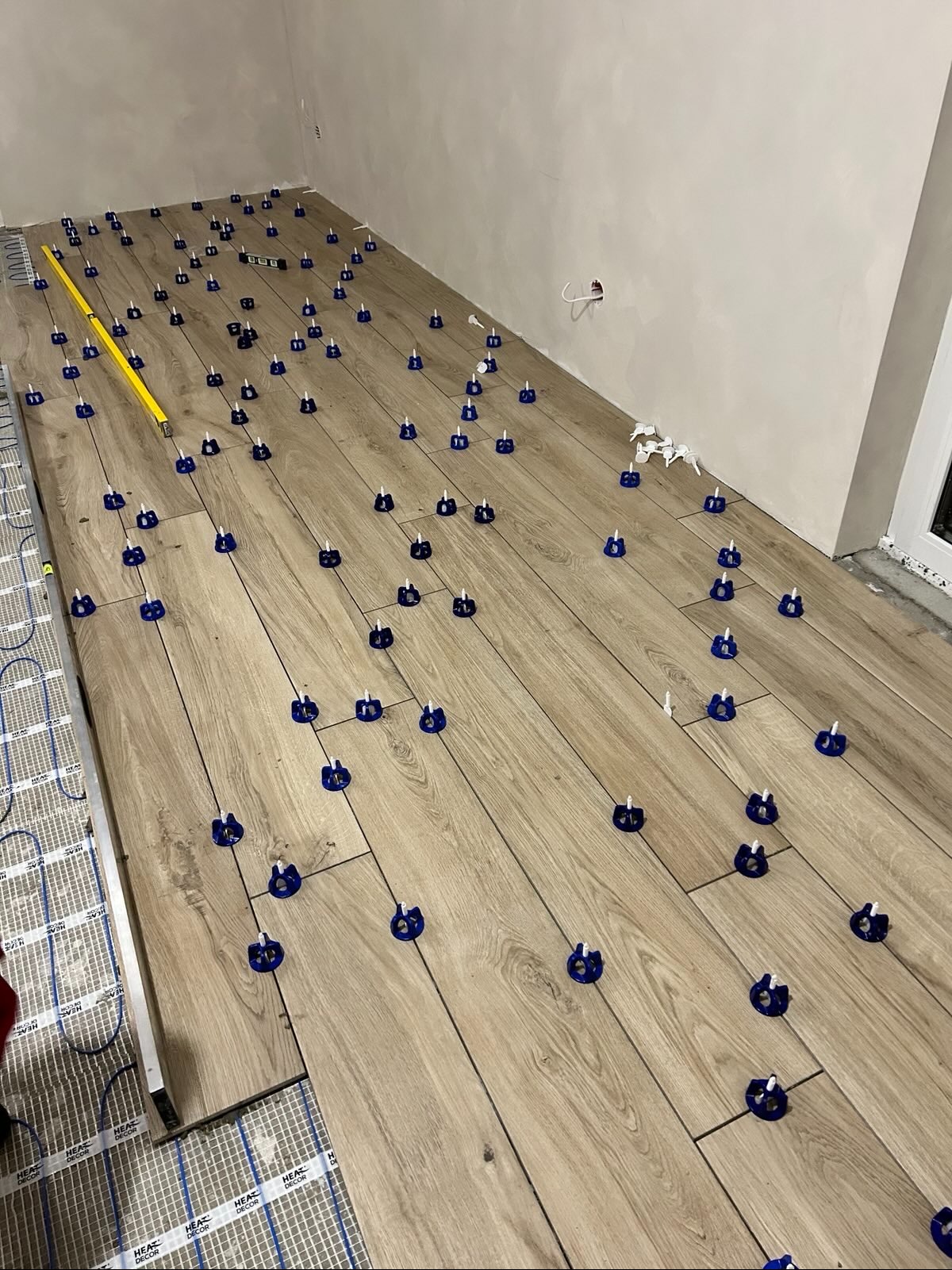 Układanie paneli podłogowych z ogrzewaniem podłogowym, z użyciem systemu poziomowania płytek. Widoczna poziomica i fragment instalacji grzewczej.