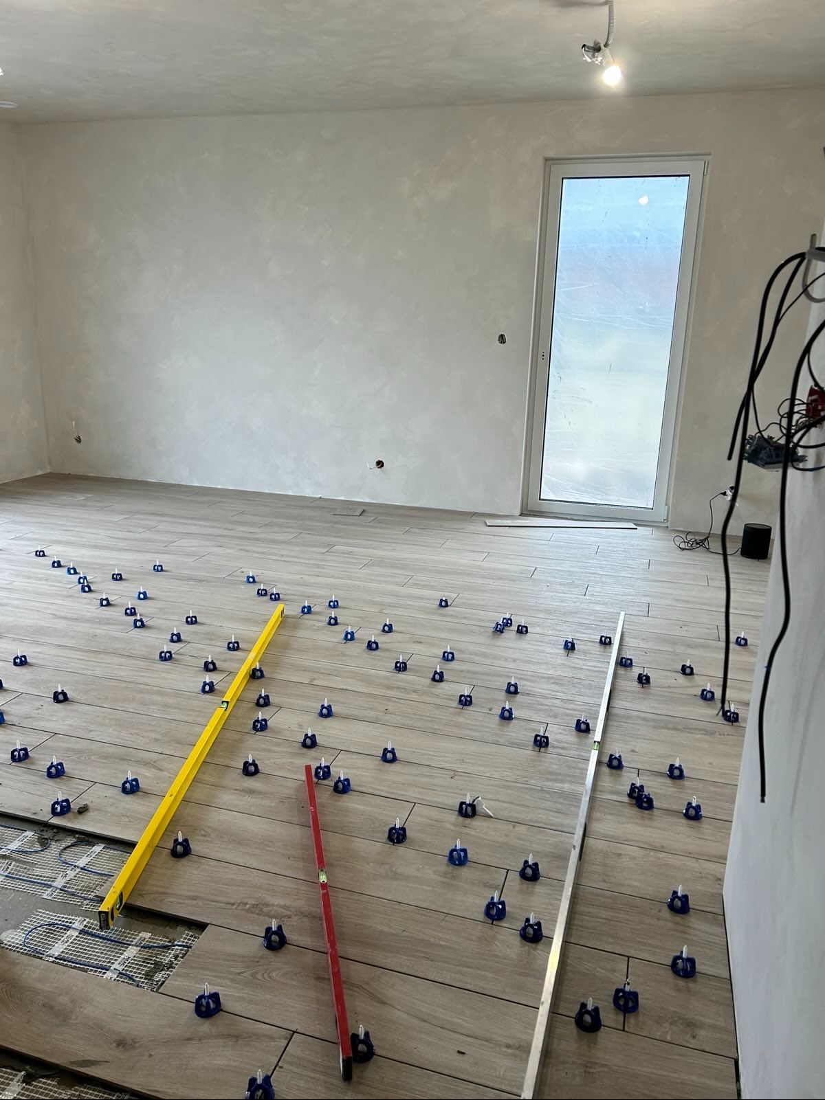 Układanie płytek podłogowych z użyciem poziomnic i klipsów w jasnym pomieszczeniu. Widoczny fragment ogrzewania podłogowego. Praca w toku, surowy stan ścian.