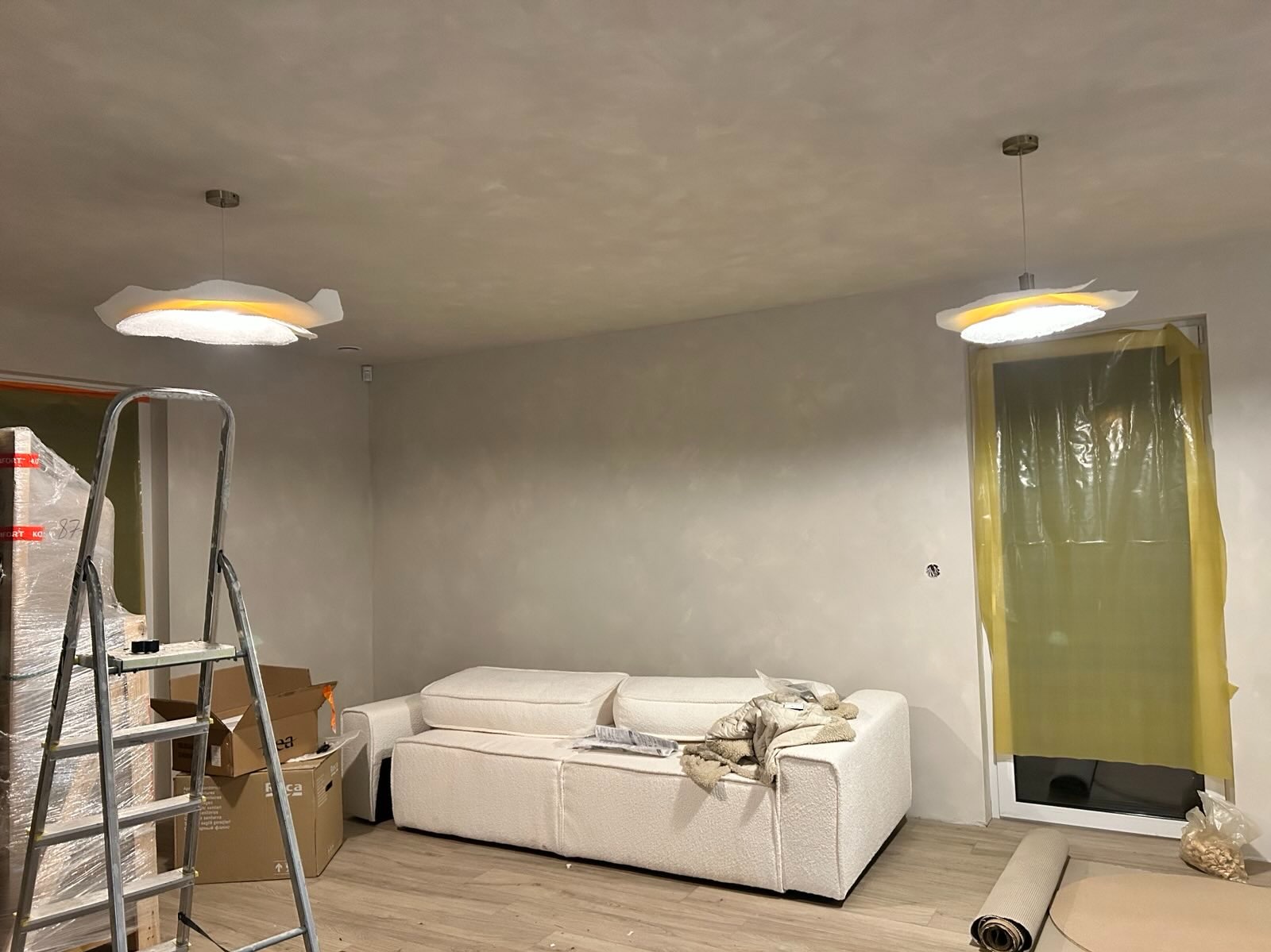 Wnętrze w trakcie wykończenia: drabina, sofa, lampy sufitowe. Ściany po gładziowaniu, okno zabezpieczone folią. Widoczne kartony i zwinięty dywan.