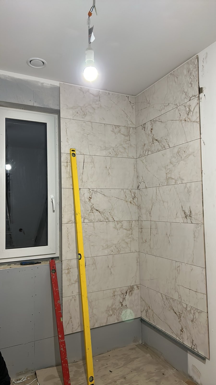 Układanie płytek imitujących marmur w rogu łazienki, widoczne poziomice: żółta i czerwona. Częściowo wykończone ściany, surowy stan podłogi. W tle okno.