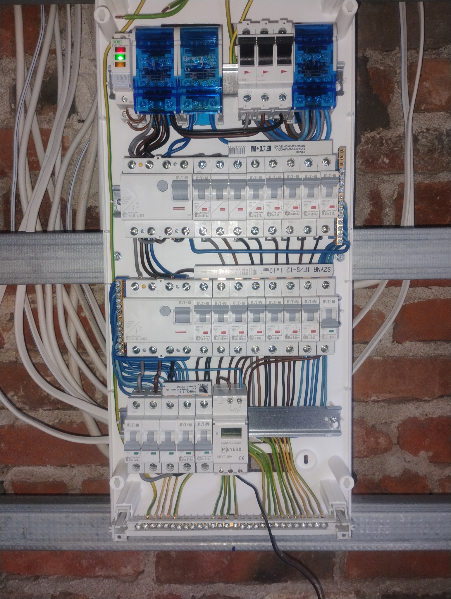 Rozdzielnica elektryczna z bezpiecznikami, przewodami i elementami sterującymi, zamontowana na ścianie z cegły. Widoczne kable i szczegóły instalacji.