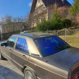 Brązowy Mercedes z przyciemnionymi na niebiesko szybami na tle domu z brązowym dachem i błękitnego nieba. Widok z tyłu, lekko z boku. Felgi aluminiowe.