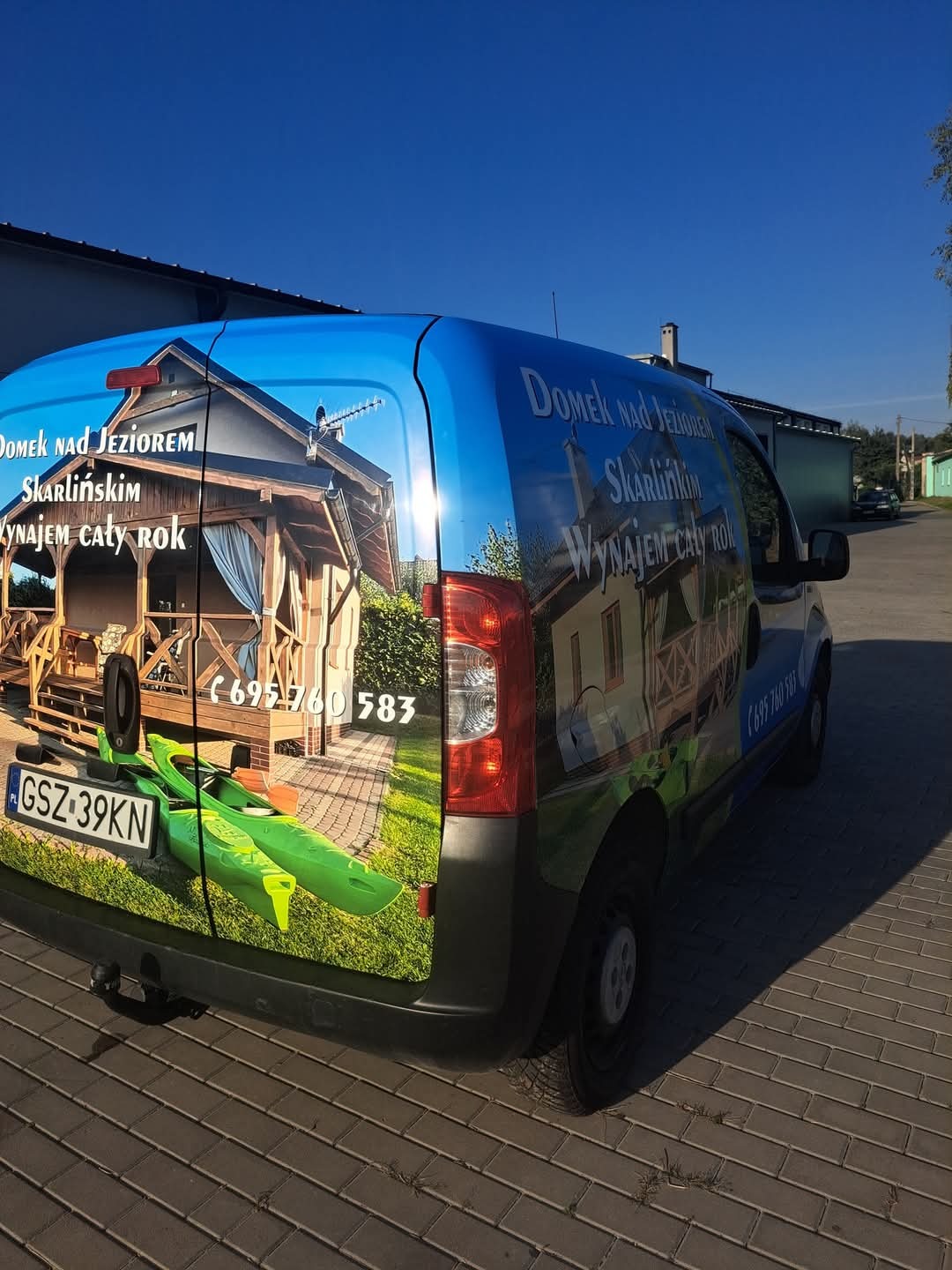 Niebieski van z oklejeniem reklamowym: domek nad jeziorem, kajak i numer telefonu. Grafika zajmuje całą tylną część pojazdu, zaparkowanego na brukowanej nawierzchni.