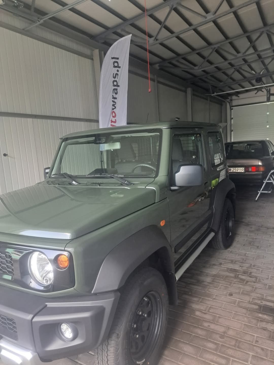 Zielony Suzuki Jimny po oklejeniu folią w Kurzętniku, widoczny w warsztacie z flagą reklamową. Detal: czarne felgi i listwy, tablica rejestracyjna innego auta w tle.