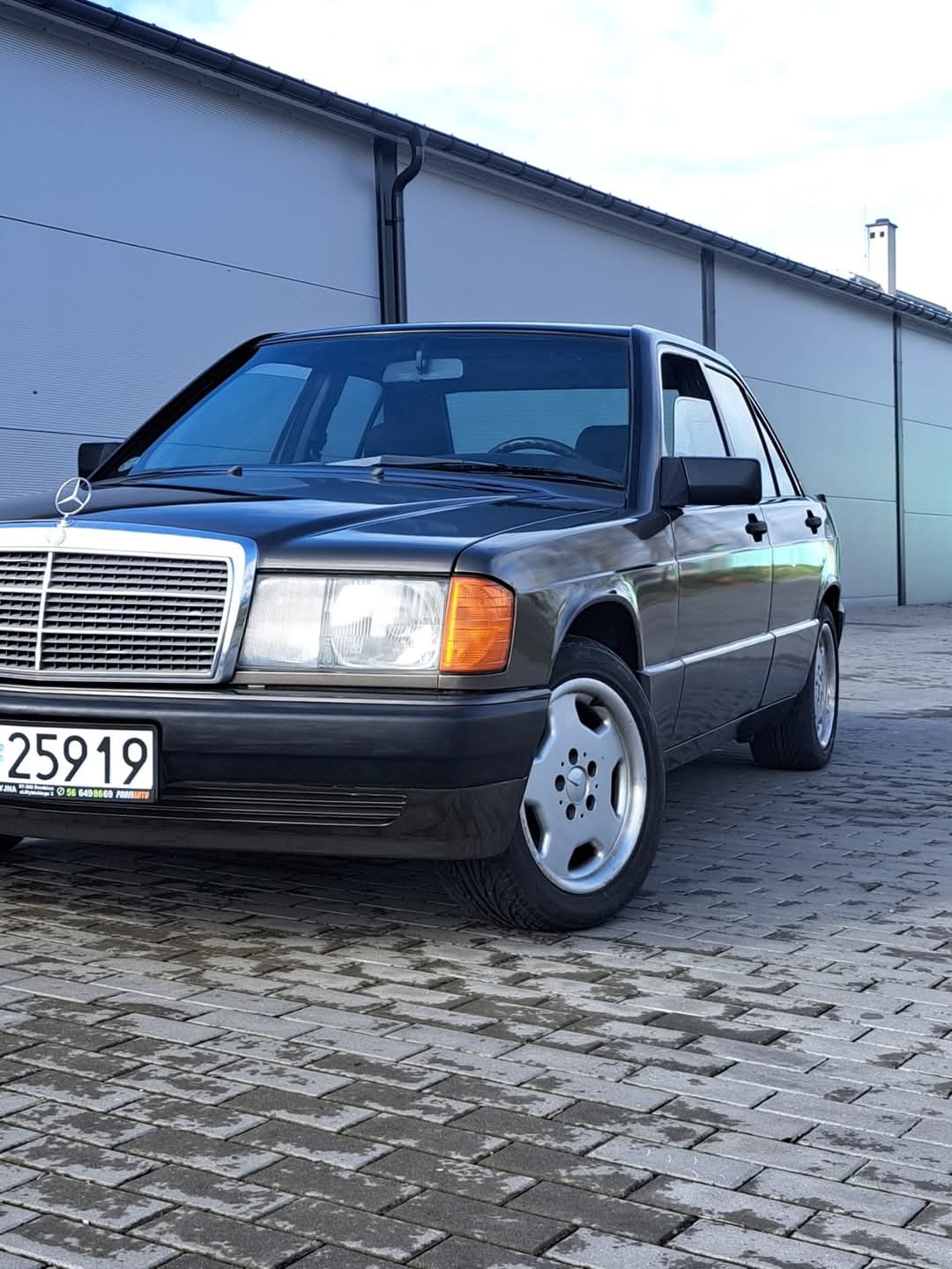 Czarny Mercedes-Benz 190E na kostce brukowej, z widoczną tablicą rejestracyjną i charakterystyczną gwiazdą na masce. Ujęcie z dołu, podkreślające klasyczny wygląd auta.