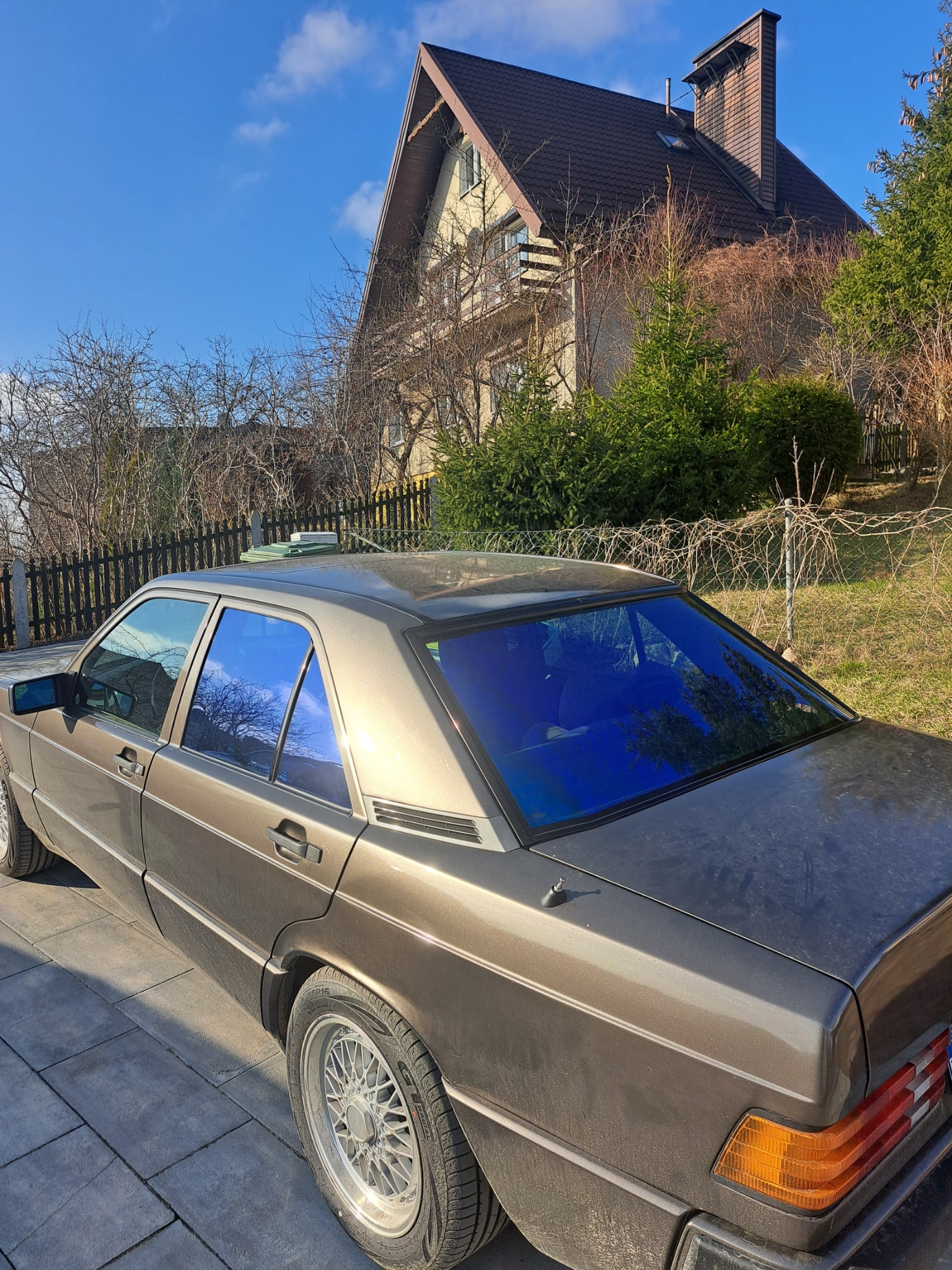 Brązowy Mercedes z przyciemnionymi na niebiesko szybami na tle domu z brązowym dachem i błękitnego nieba. Widok z tyłu, lekko z boku. Felgi aluminiowe.