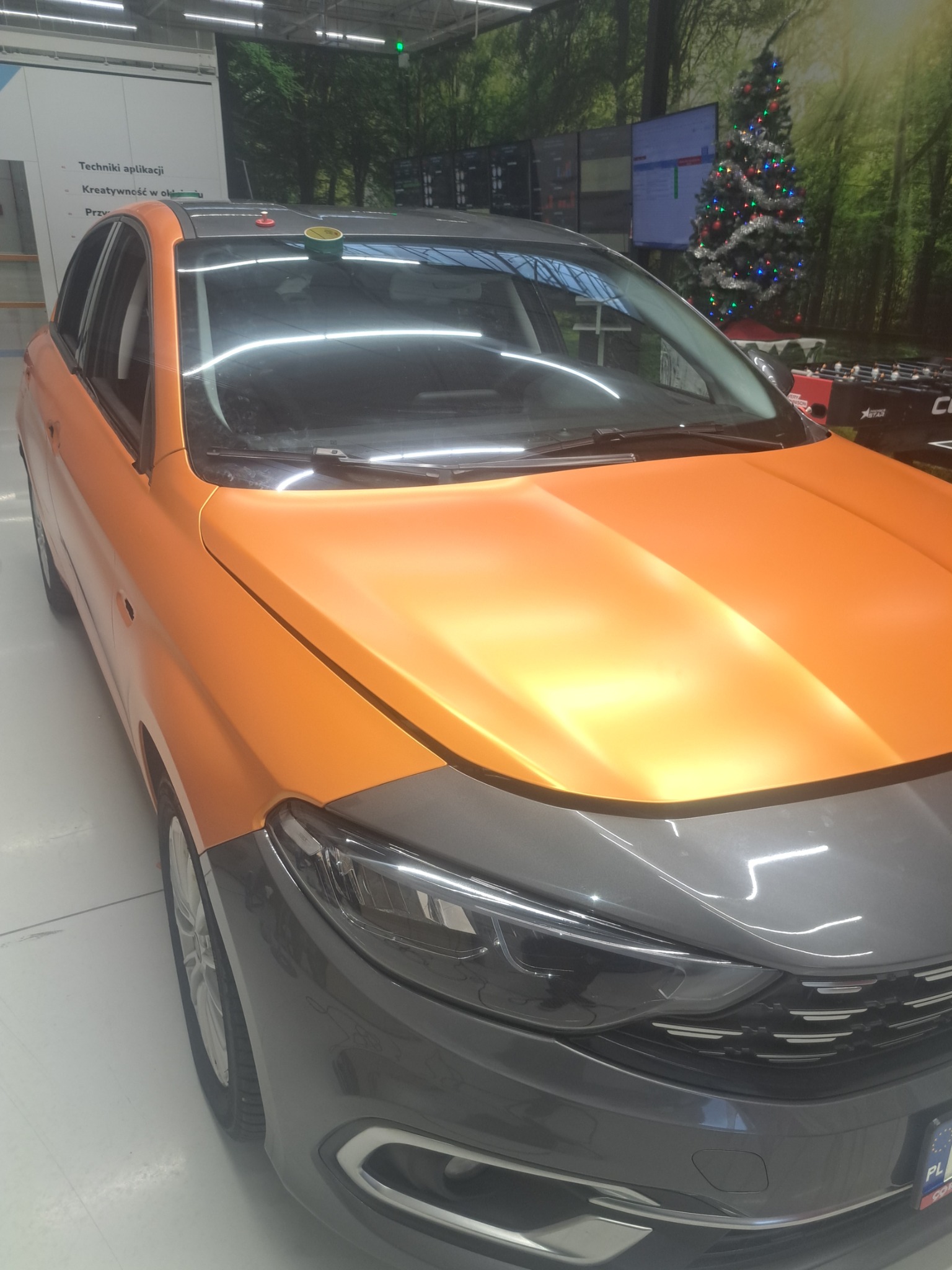 Oklejony na pomarańczowo samochód z szarym przodem, detale oklejenia zabezpieczone taśmą. W tle choinka i grafika lasu.