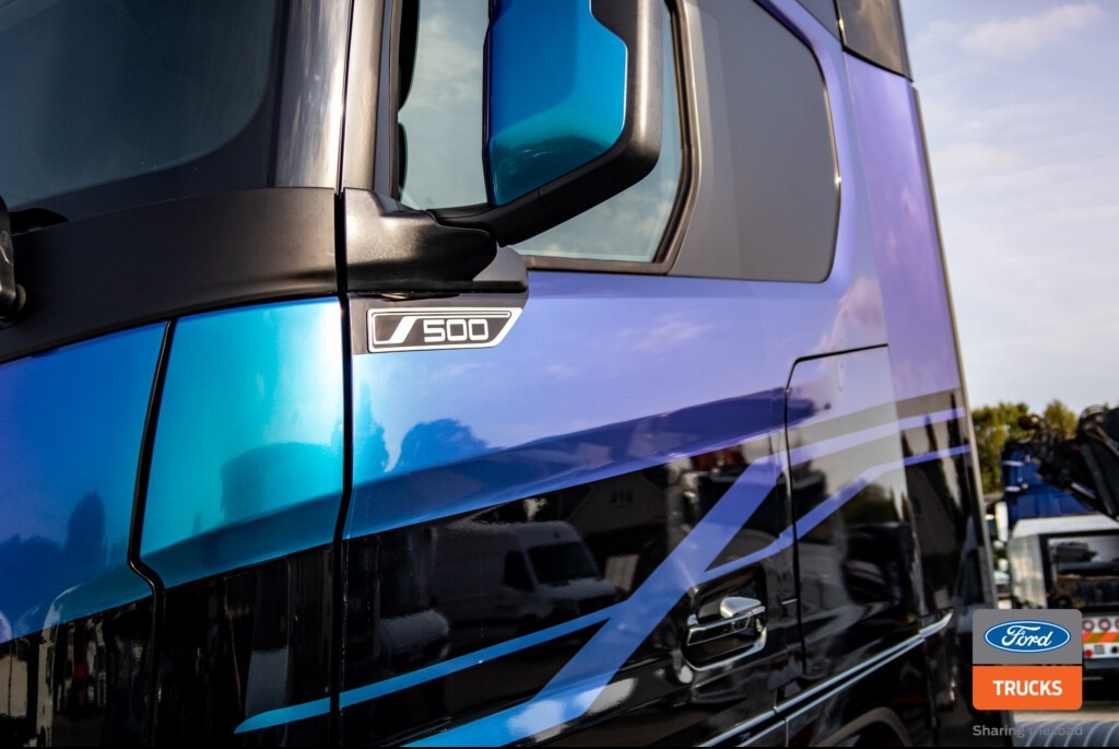 Błyszcząca, niebiesko-fioletowa okleina na kabinie ciężarówki Ford Trucks S500 z czarnymi akcentami i logo marki w rogu. Widoczne odbicia otoczenia.