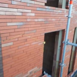 Perfect Home Remonty i wykończenia wnętrz - Murowana ściana budynku z czerwonej cegły, widoczne okno i otwory drzwiowe, poziomica sznurkowa, fragment rusztowania. Prace murarskie w toku.