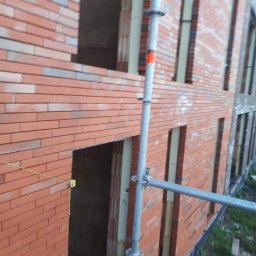 Perfect Home Remonty i wykończenia wnętrz - Wznoszenie muru z cegły klinkierowej na budynku w trakcie budowy. Widoczne rusztowanie i otwory okienne. Częściowo wykończona fasada budynku.