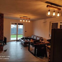 Perfect Home Remonty i wykończenia wnętrz - Jasne wnętrze salonu z ceglanym kominkiem i nowoczesnym oświetleniem. Widoczne meble i okno z widokiem na ogród. Minimalistyczny wystrój.