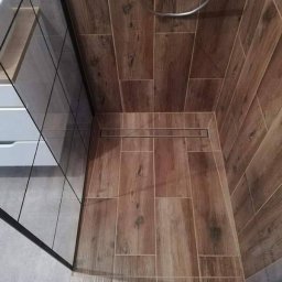 Perfect Home Remonty i wykończenia wnętrz - Wykończona kabina prysznicowa z drewnopodobnymi płytkami na ścianach i podłodze, z odpływem liniowym. Widoczna szklana ściana kabiny i fragment szafki umywalkowej.