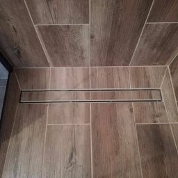 Perfect Home Remonty i wykończenia wnętrz - Ukończona kabina prysznicowa z drewnopodobnymi płytkami i odpływem liniowym. Widok z góry, podkreślający precyzję ułożenia płytek i estetykę wykończenia.