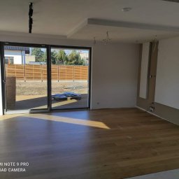 Perfect Home Remonty i wykończenia wnętrz - Jasne wnętrze z parkietem i dużym oknem przesuwnym. Widoczne niedokończone prace elektryczne i fragment zabudowy na ścianie. Na zewnątrz drewniany płot.