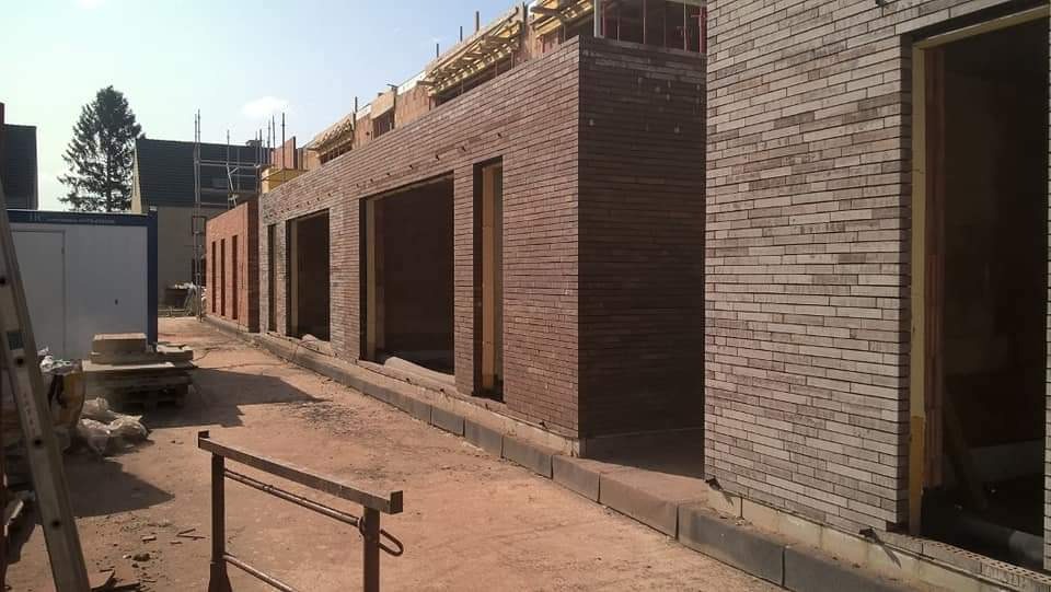 Budynek w trakcie budowy z cegły, widoczne otwory okienne i drzwiowe, elementy konstrukcji dachowej, kontener budowlany w tle.