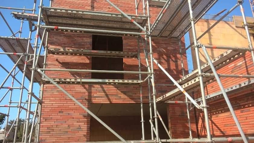 Wznoszenie dwukondygnacyjnego budynku z czerwonej cegły, widoczne rusztowania i otwory okienne. Budowa w toku, widok z dołu na fragment ściany i niebieskie niebo.