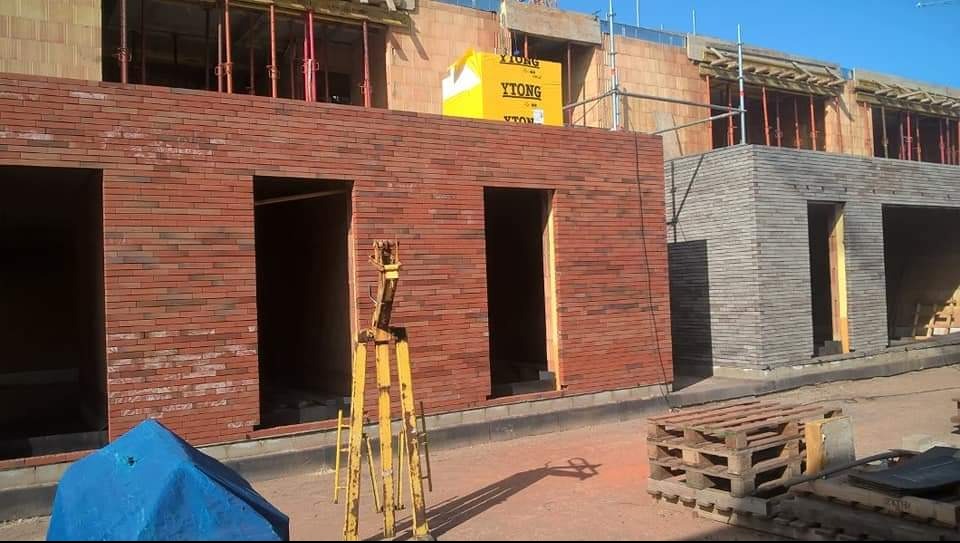 Murowanie ścian budynku z czerwonej cegły. Widoczne otwory okienne i drzwiowe. Na górze bloki YTONG. Na pierwszym planie palety i statyw geodezyjny.