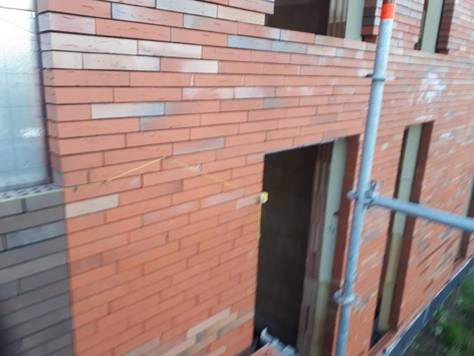 Murowana ściana budynku z czerwonej cegły, widoczne okno i otwory drzwiowe, poziomica sznurkowa, fragment rusztowania. Prace murarskie w toku.