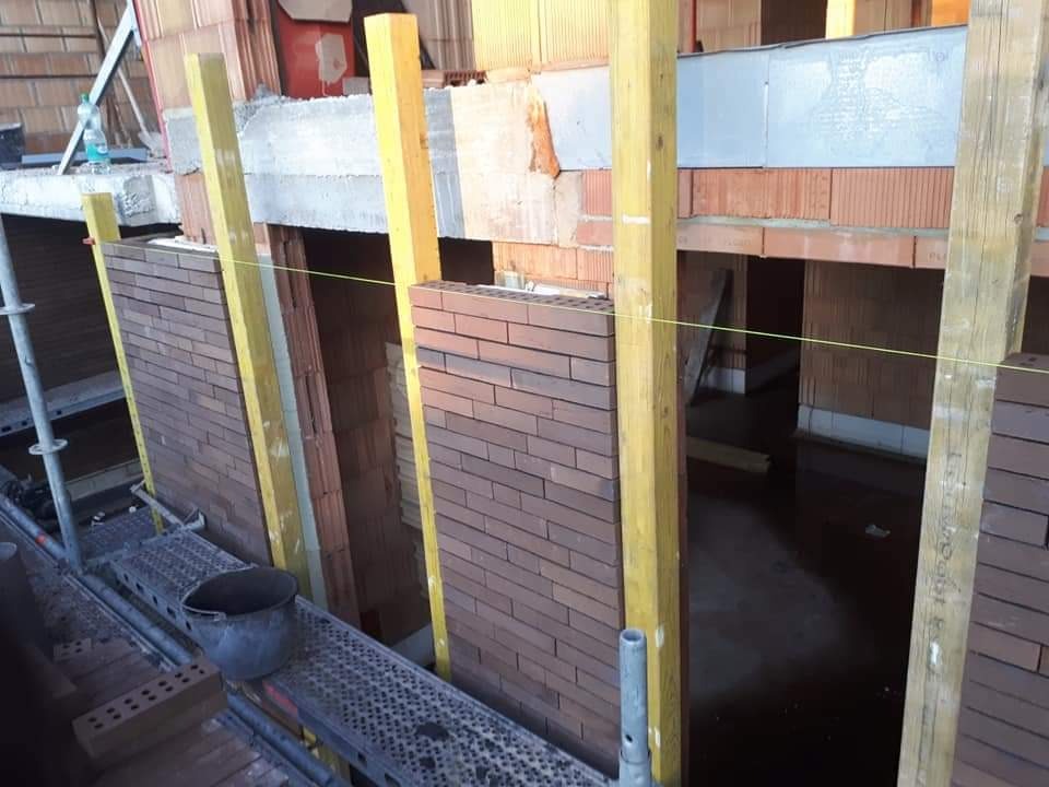 Murowanie ściany z cegły klinkierowej na budowie. Widoczne rusztowanie, podpory i sznurek murarski. W tle ściany z pustaków ceramicznych.
