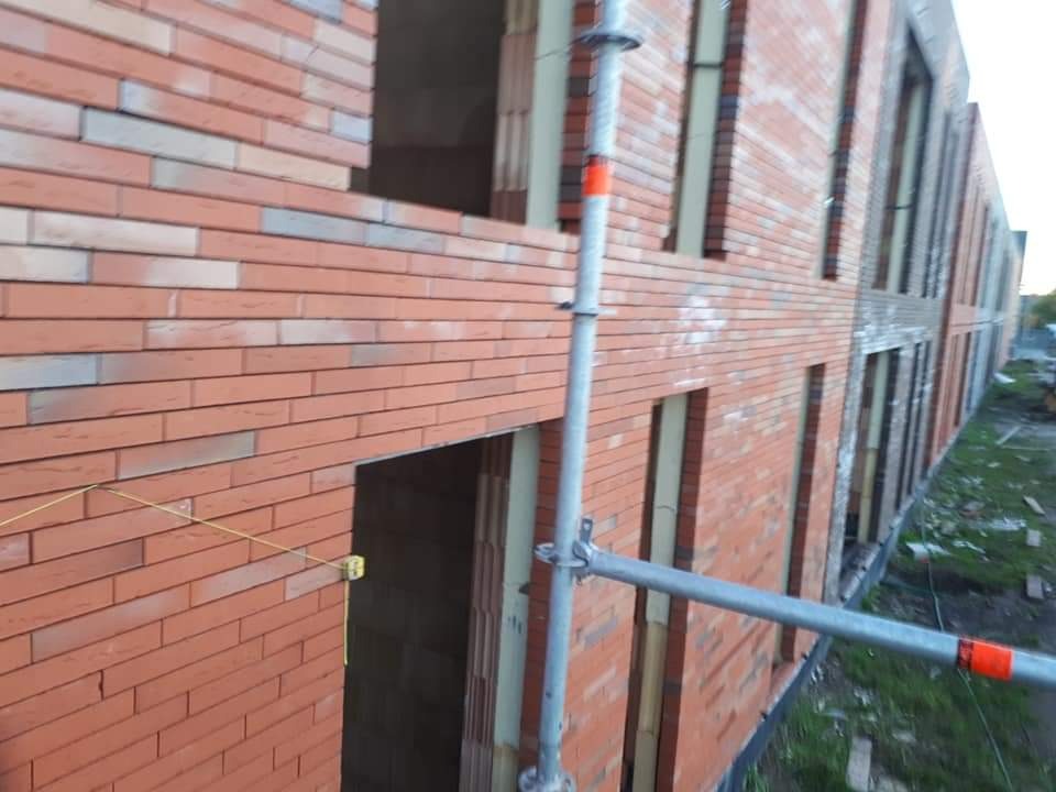 Wznoszenie muru z cegły klinkierowej na budynku w trakcie budowy. Widoczne rusztowanie i otwory okienne. Częściowo wykończona fasada budynku.