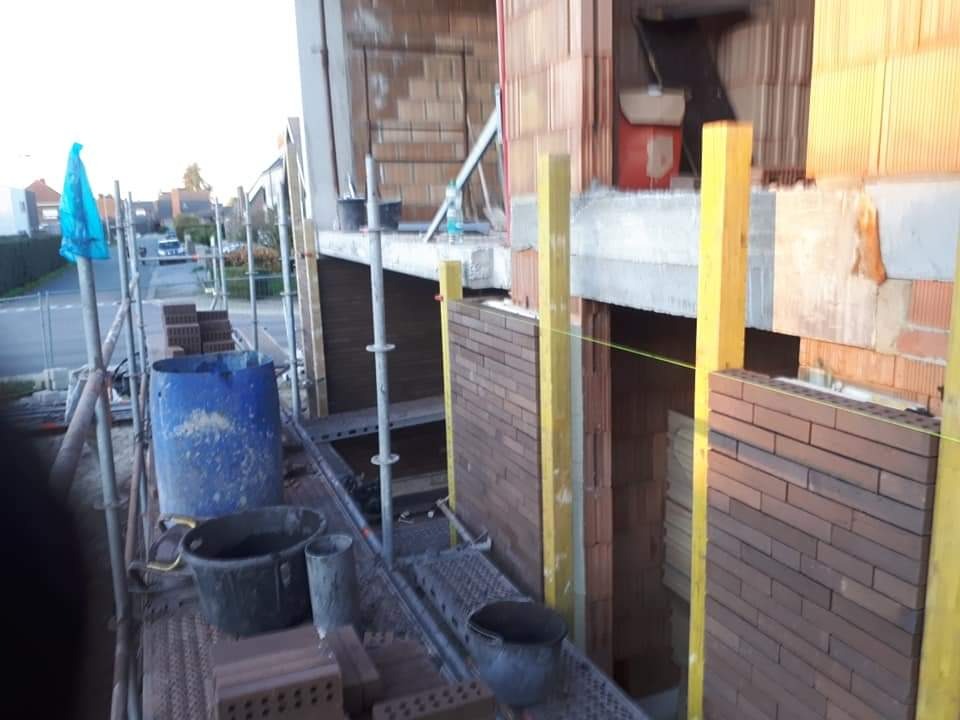 Murowanie ścian zewnętrznych z cegły na rusztowaniu, widok z góry na plac budowy z narzędziami i materiałami budowlanymi.