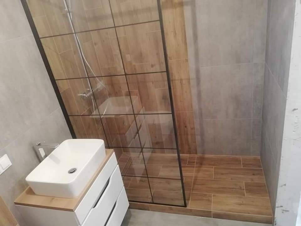 Nowoczesna łazienka z kabiną prysznicową typu walk-in, umywalką nablatową i szarymi płytkami. Wnętrze wykończone w ciepłych odcieniach drewna.
