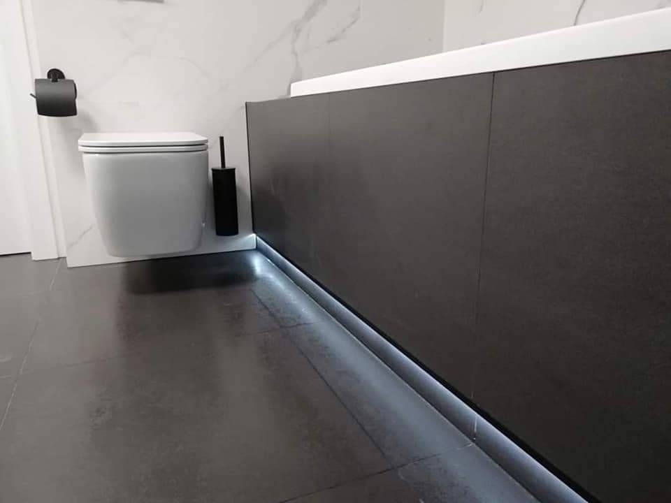 Nowoczesna łazienka z podświetleniem LED wzdłuż wanny, szara glazura podłogowa, podwieszana toaleta i marmurowe wykończenie ściany.