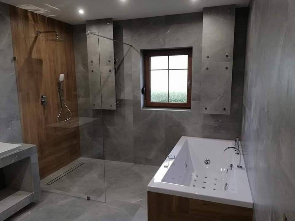 Nowoczesna łazienka z kabiną prysznicową typu walk-in, wanną z hydromasażem i oknem. Szare płytki imitujące marmur, drewniane akcenty i minimalistyczne oświetlenie.