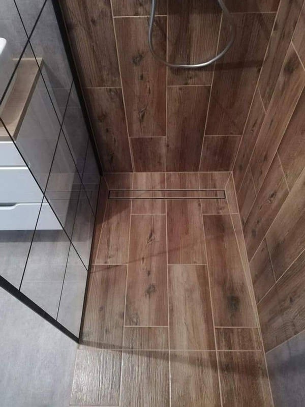 Wykończona kabina prysznicowa z drewnopodobnymi płytkami na ścianach i podłodze, z odpływem liniowym. Widoczna szklana ściana kabiny i fragment szafki umywalkowej.