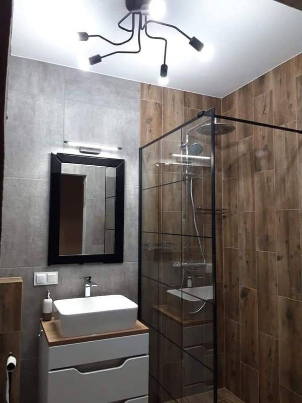 Nowoczesna łazienka z kabiną prysznicową typu walk-in, umywalką nablatową i oryginalną lampą sufitową. Płytki imitujące drewno i szarość tworzą spójną całość.
