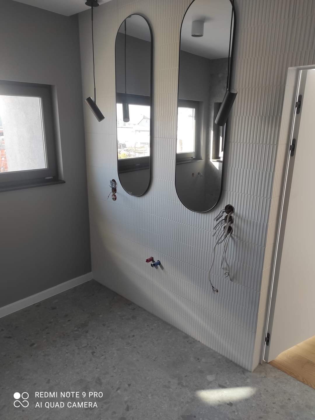 Szara łazienka w trakcie wykończenia: owalne lustra, płytki z fakturą, widoczne instalacje, surowy beton na podłodze i minimalistyczne oświetlenie.