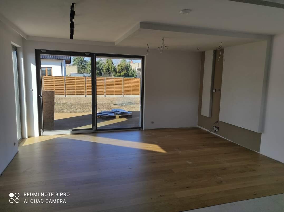 Jasne wnętrze z parkietem i dużym oknem przesuwnym. Widoczne niedokończone prace elektryczne i fragment zabudowy na ścianie. Na zewnątrz drewniany płot.