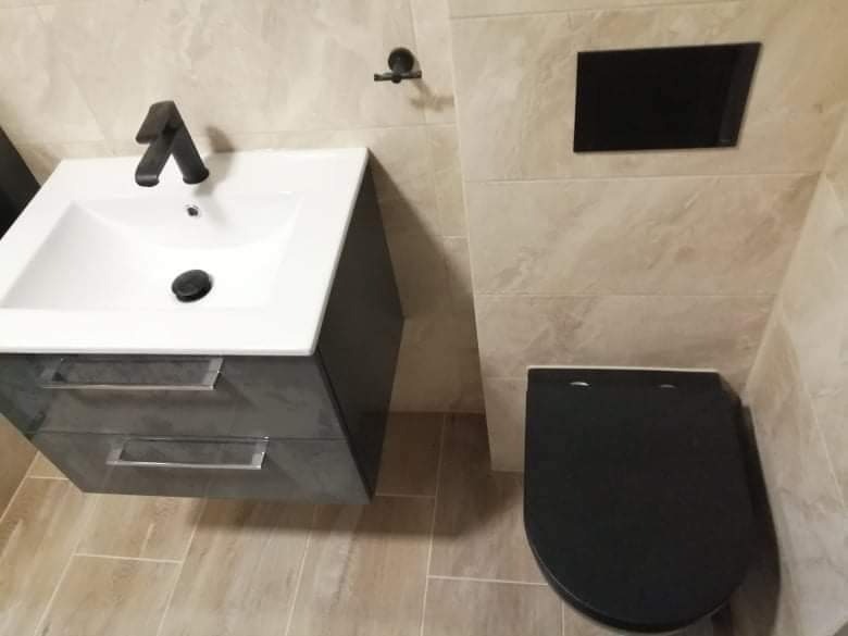 Umywalka z szafką, toaleta z czarną deską i beżowe płytki w wykończonej łazience. Minimalistyczne wnętrze z ciemnymi akcentami.